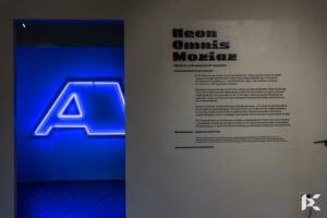 Zdjęcie z wystawy neonów - na pierwszym planie widać tekst wprowadzający do wystawy Neon Omnis Moriar, w tle neon z literą A z napisu Wólczanka.