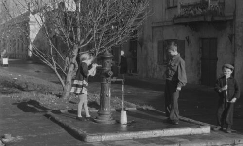 Czarni biała fotografia ukazująca dziewczynkę pompującą wodę do kanki, obok starszy chłopiec, w tle budynki