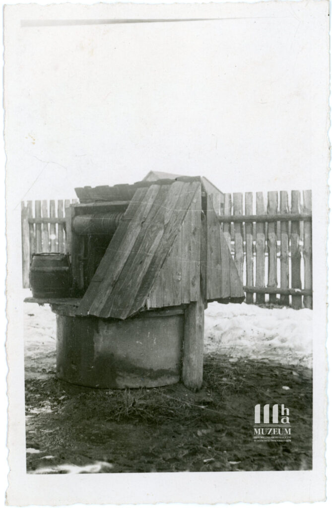 Fotografia czarno-biała przedstawiająca studnię z betonowego kręgu z trójkątną zamykaną drewnianą nadbudówką, w tle widoczny fragment drewnianego płotu oraz wydeptany śnieg