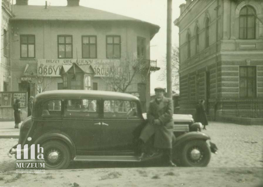 Fotografia czarno biała mężczyzny opierającego się o samochód, w tle budynek kina Amida, na elewacji zapowiedź filmu Ordynat Michorowski z 1937 r. 