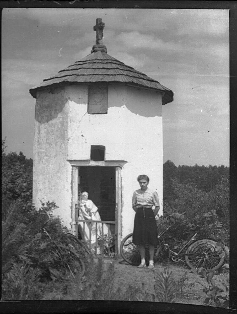 Fotografia czarno-biała, w centralnej części zdjęcia kaplica na tle niewielkich drzew, w wejściu oparta rzeźba św.. Katarzyny, na zewnątrz przed wejściem dziewczyna w spódnicy i bluzce, obok stoją oparte o krzaki rowery