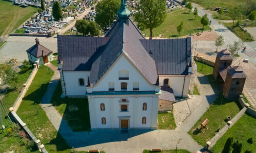 Kościół pw. Wniebowzięcia Najświętszej Maryi Panny w Krynkach widziany z lotu ptaka - fotografia barwna ukazująca budynek na planie krzyża z widoczną dzwonnicą- sygnaturką na dachu. Kościół leży na wzgórzu otoczonym murkiem, w tle widać cmentarz