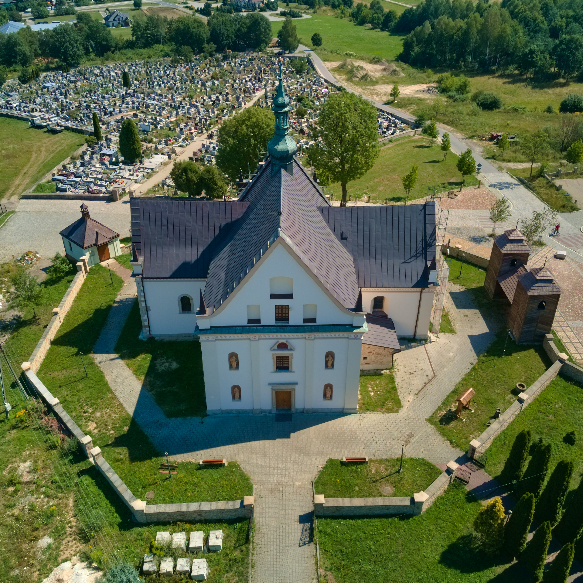 Kościół pw. Wniebowzięcia Najświętszej Maryi Panny w Krynkach widziany z lotu ptaka - fotografia barwna ukazująca budynek na planie krzyża z widoczną dzwonnicą- sygnaturką na dachu. Kościół leży na wzgórzu otoczonym murkiem, w tle widać cmentarz