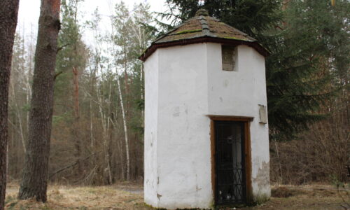 Fotografia przedstawiająca kaplicę na tle lasu - niewielki ośmioboczny budynek kryty takimż gontem, widoczne wejście i herb Dębno nad nim