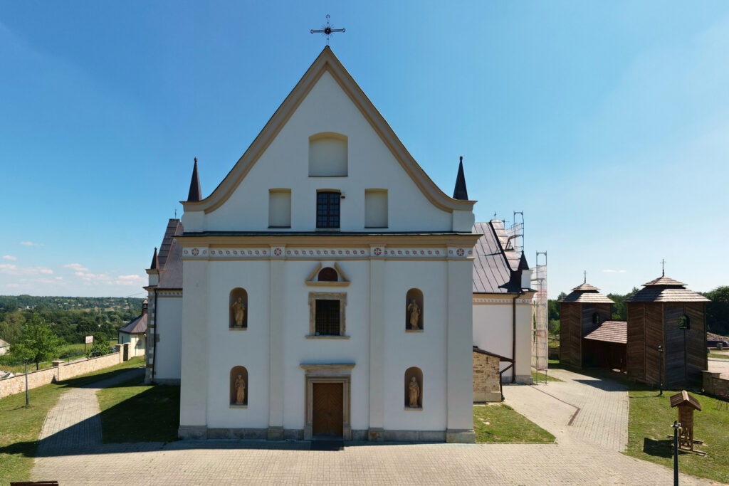Fotografia barwna przedstawiająca fasadę kościoła pw. Wniebowzięcia NMP, dwukondygnacyjną trzyosiową trójkątnie zwieńczoną