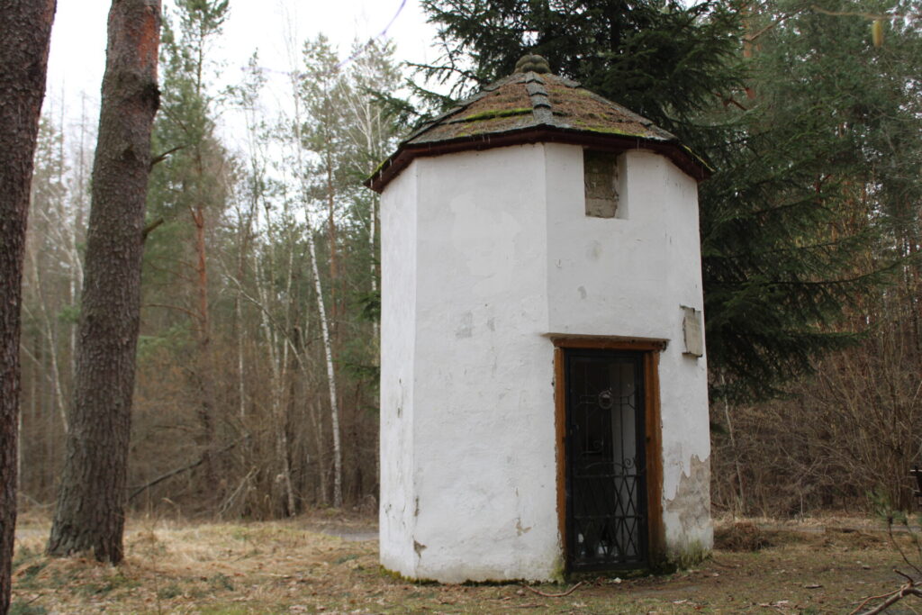 Fotografia przedstawiająca kaplicę na tle lasu - niewielki ośmioboczny budynek kryty takimż gontem, widoczne wejście i herb Dębno nad nim