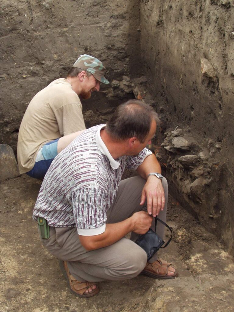 Fotografia przestawiająca dwóch archeologów w czasie pracy - obaj kucają przy ziemnym wykopie