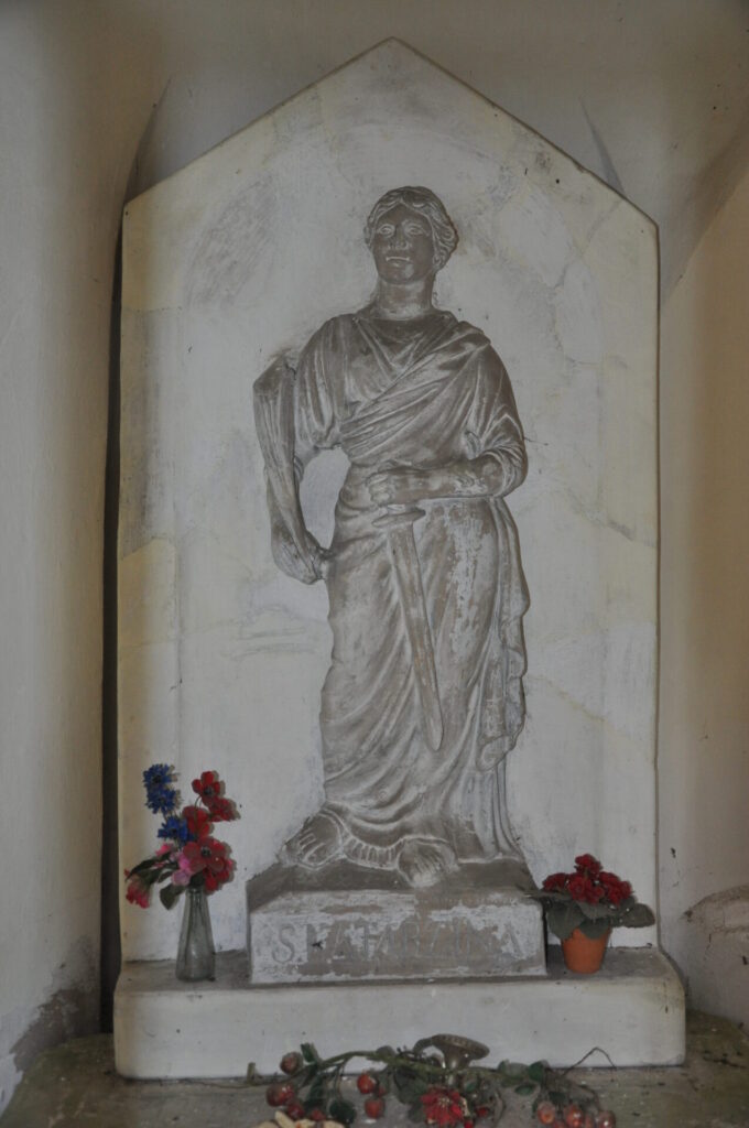 Płaskorzeźbiona figura Św. Katarzyny Aleksandryjskiej ukazanej w stroju rzymskiej kobiety - w udrapowanej szacie, z mieczem w ręce oraz gałązką palmy w drugiej. Rzeźba stanowi całość z trójkątnie zwieńczonym tłem za nią. Na bazie pod stopami podpis S. Katarzina