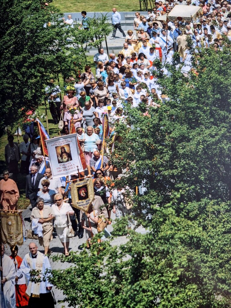 Fotografia przedstawia procesję Bożego Ciała w Ostrowcu Świętokrzyskim - widać tłum ludzi z chorągwiami, idących między drzewami. Zdjęcie wykonano z góry.