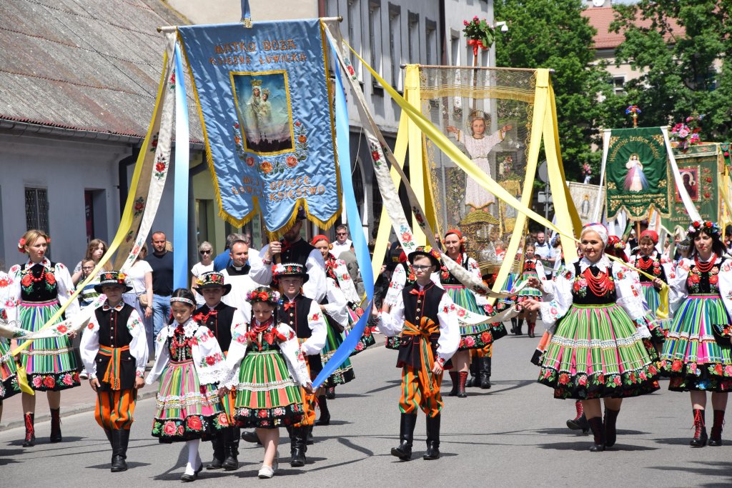 Zdjęcie współczesne przedstawiające procesję Bożego Ciała - zdjęcie ukazuje dzieci ubrane w stroje łowickie - koszule i pasiaste spódnice, niosące chorągwie ze wstęgami