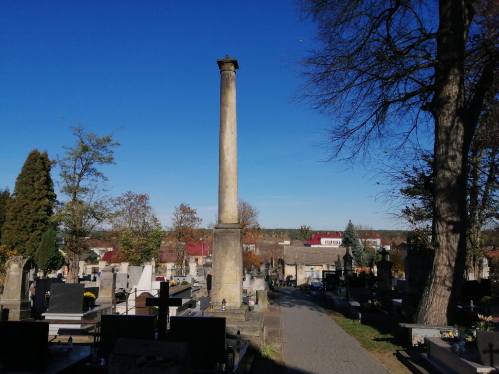 Zdjęcie przedstawiające kolumnę stojącą na cmentarzu w Ćmielowie - wysoki obelisk na prostopadłościennej podstawie.