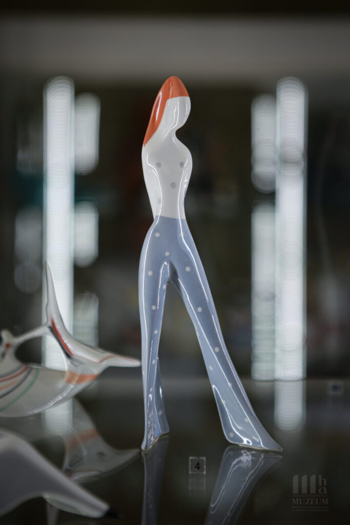 Porcelanowa figurka - dziewczyna stojąca z jedną nogą wysuniętą. patrzy w lewą stronę, włosy rudego koloru ma związane w wysoki kucyk, dłonie trzyma za plecami. Ubrana jest w niebieskie spodnie w białe groszki, groszki tego samego koloru co spodnie ma na białej górnej części tułowia. Figurka o bardzo schematycznie ukazanych kształatach.